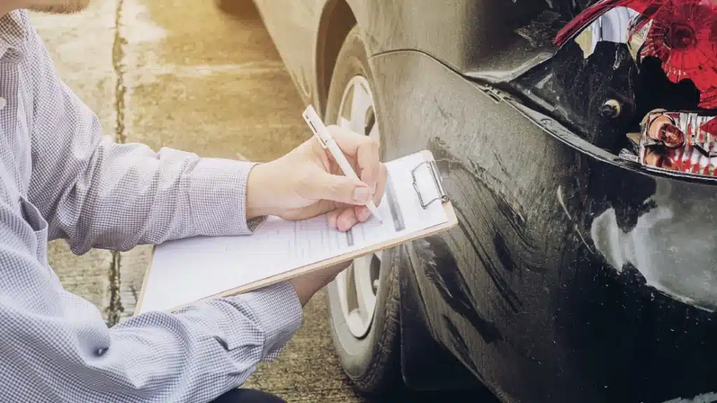 Foto de uma pessoa checando algo específico em um carro batido e realizando anotações em alusão a pauta "o que é veículo sinistrado"