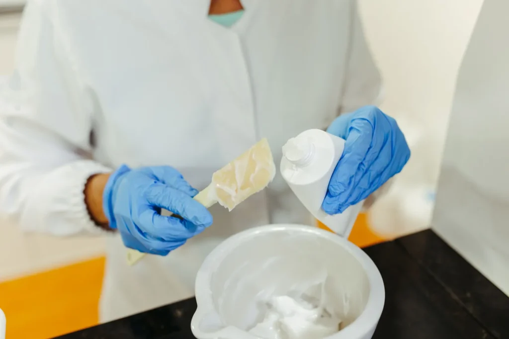 Profissional com jaleco branco e luvas azuis despeja creme de um tubo em um recipiente.