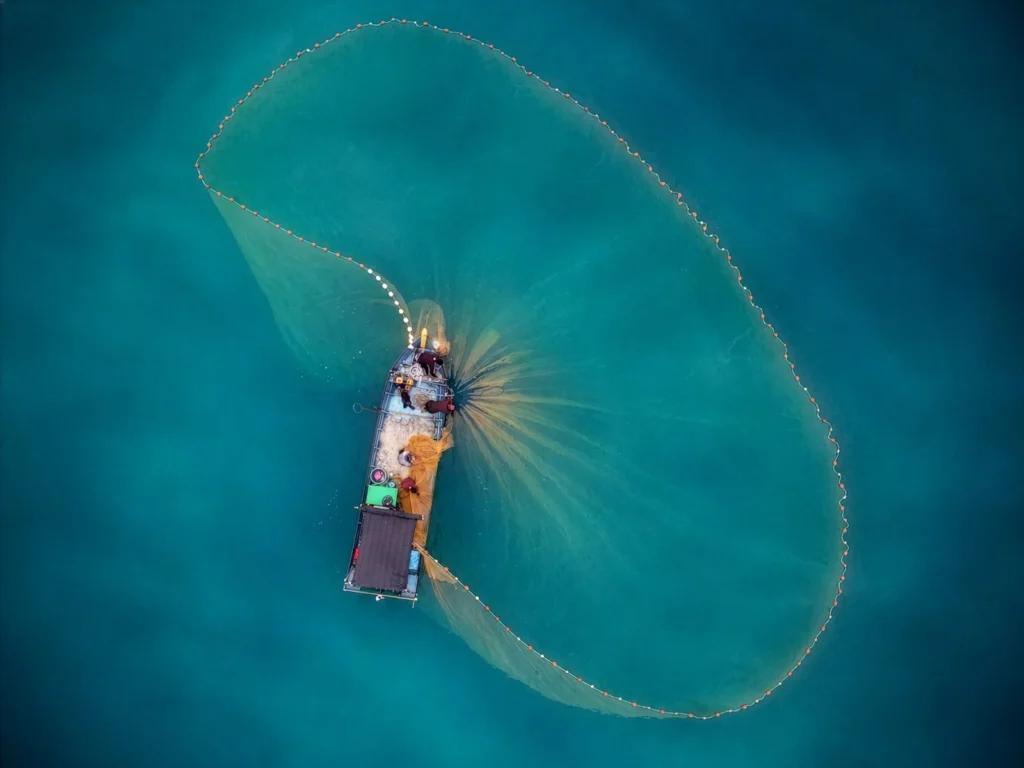 Vista aérea de barco de pesca cercando a água com uma rede circular verde.