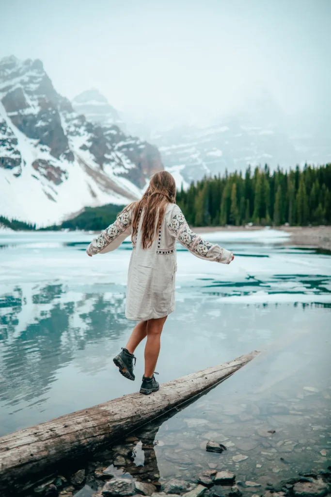 Mulher de costas com vestido branco e botas caminhando sobre tronco em lago alpino