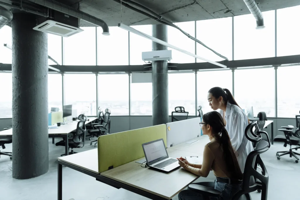 Duas mulheres trabalhando em computadores em um escritório corporativo amplo e iluminado.