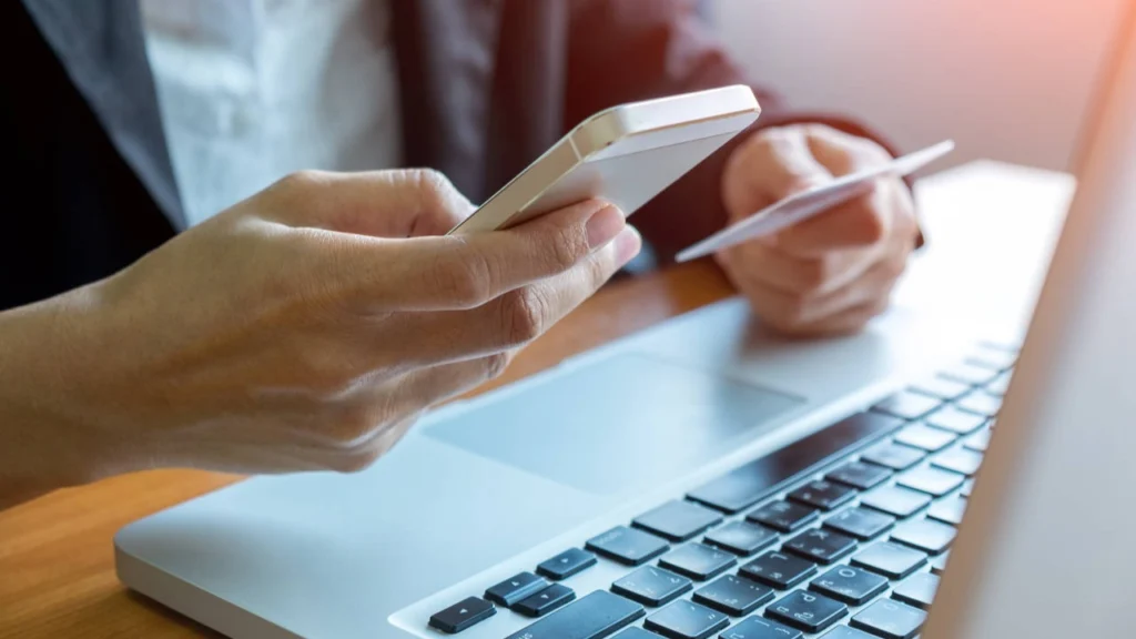 Imagem atual: Foto de uma pessoa consultando a CNH pelo celular com documentos em mãos e um notebook em frente em alusão a pauta sobre "Como acessar CNH Digital"